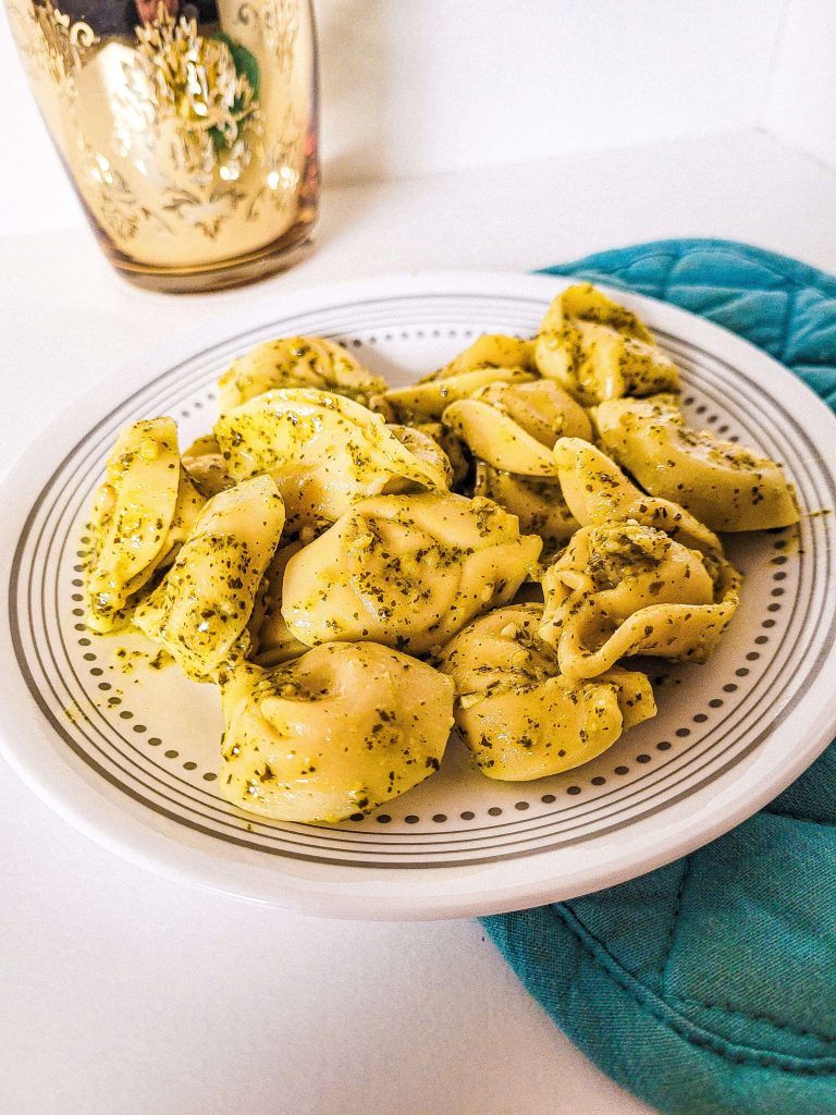 pesto tortellini on a plate with a gold cu in the background. 