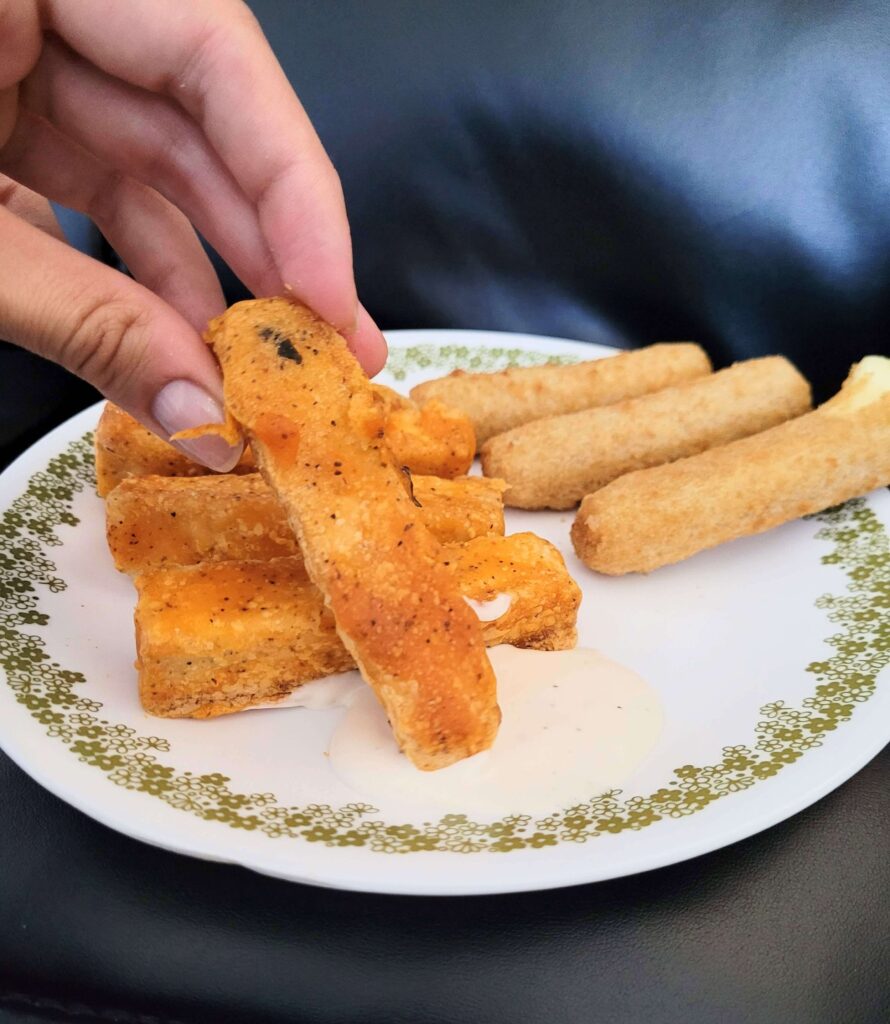 buffalo tofu with a hand holding up a piece on a plate with mozzarella sticks in the background.