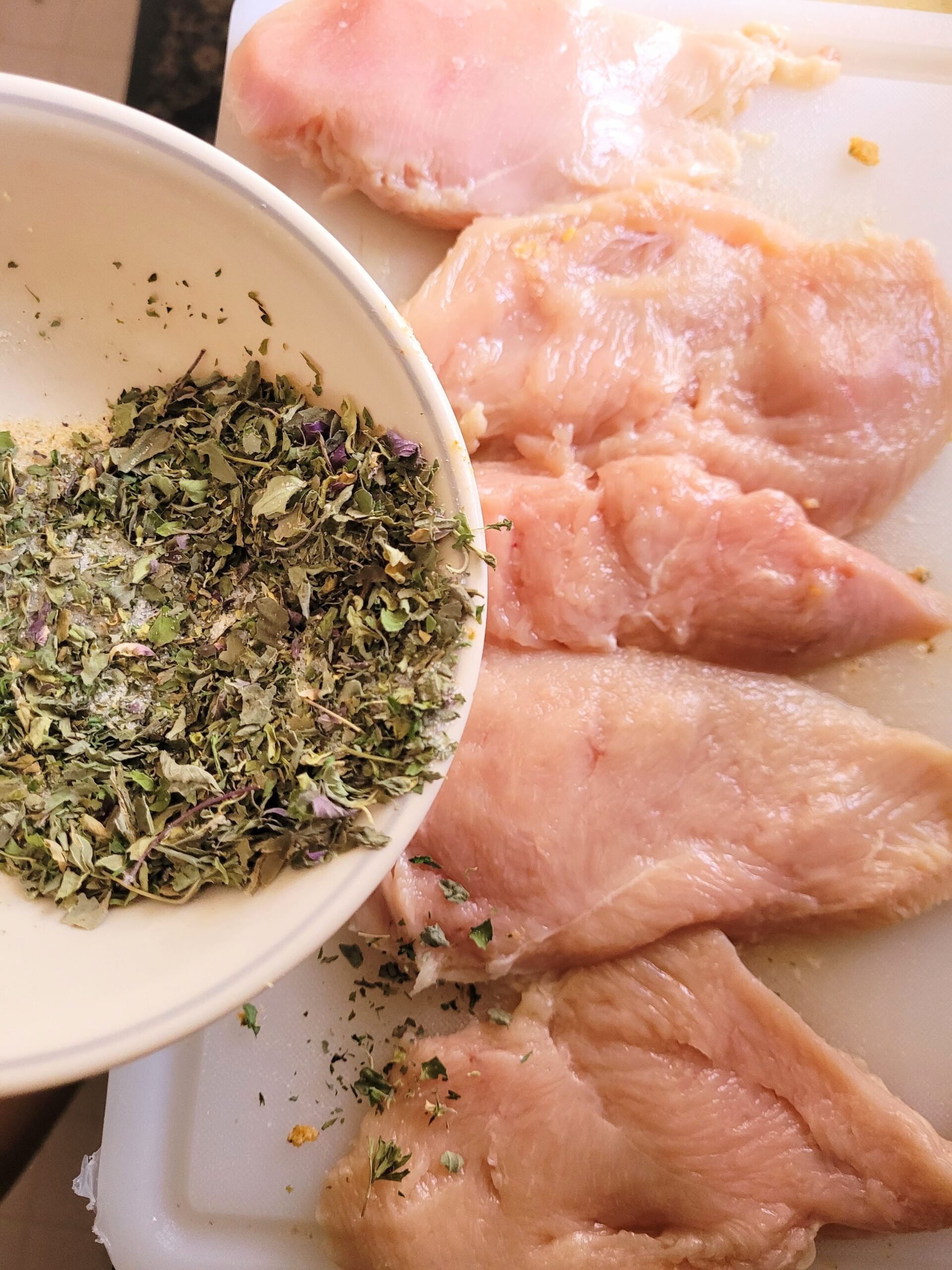 herb seasoning in bowl being sprinkled on top chicken breast.