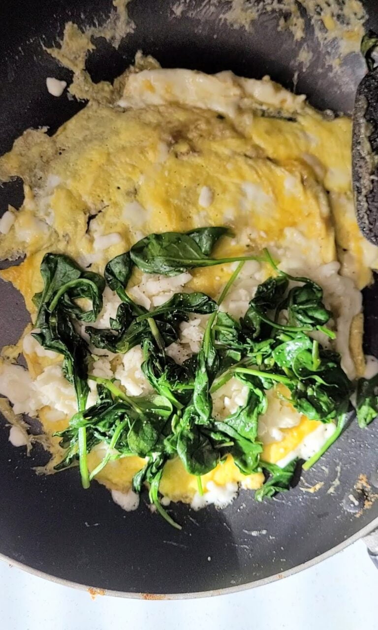 Omelet frying in skillet with spinach and cheese on top.